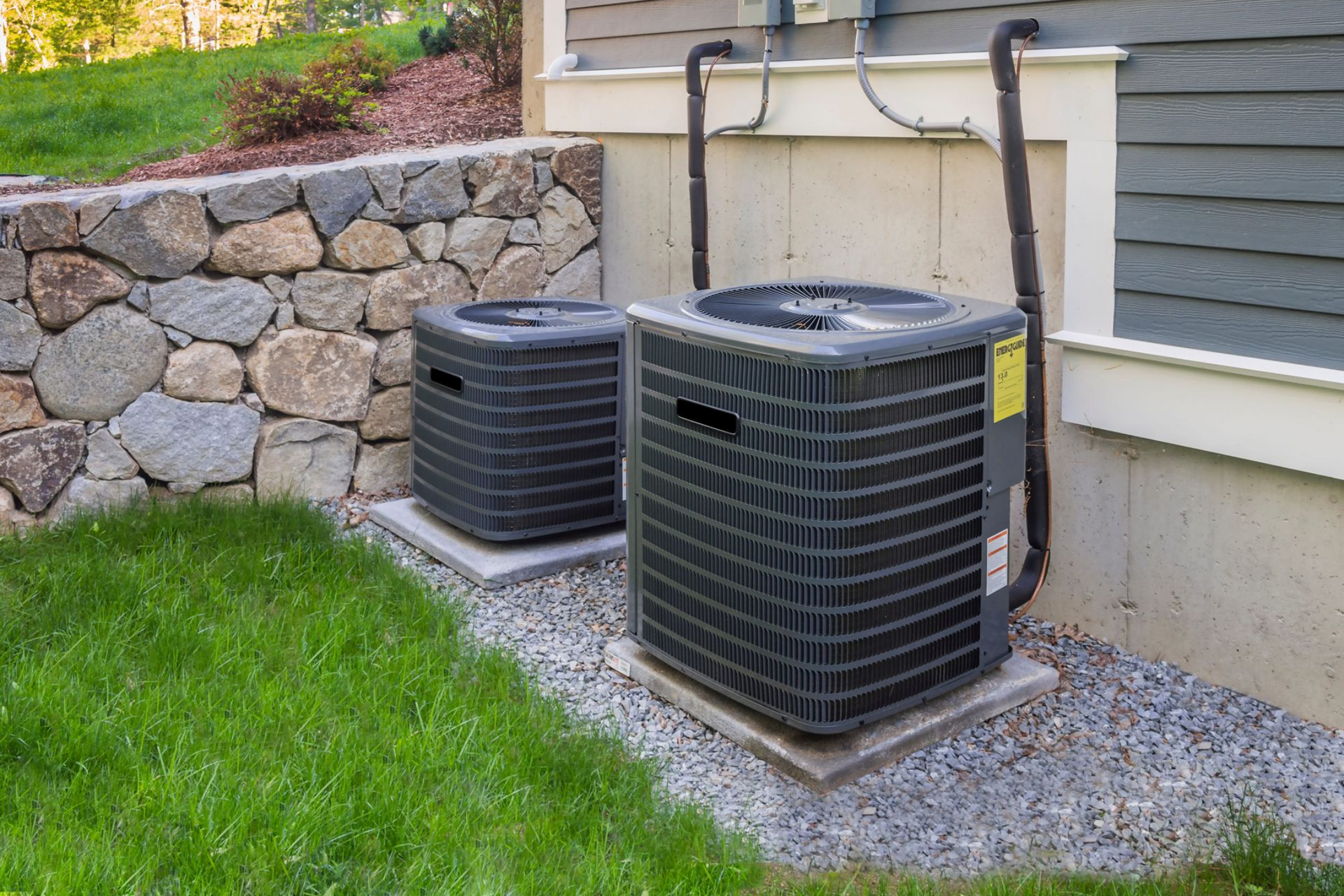 Two gray HVAC condenser units sit on gravel pads next to a house wall and a stone retaining wall.