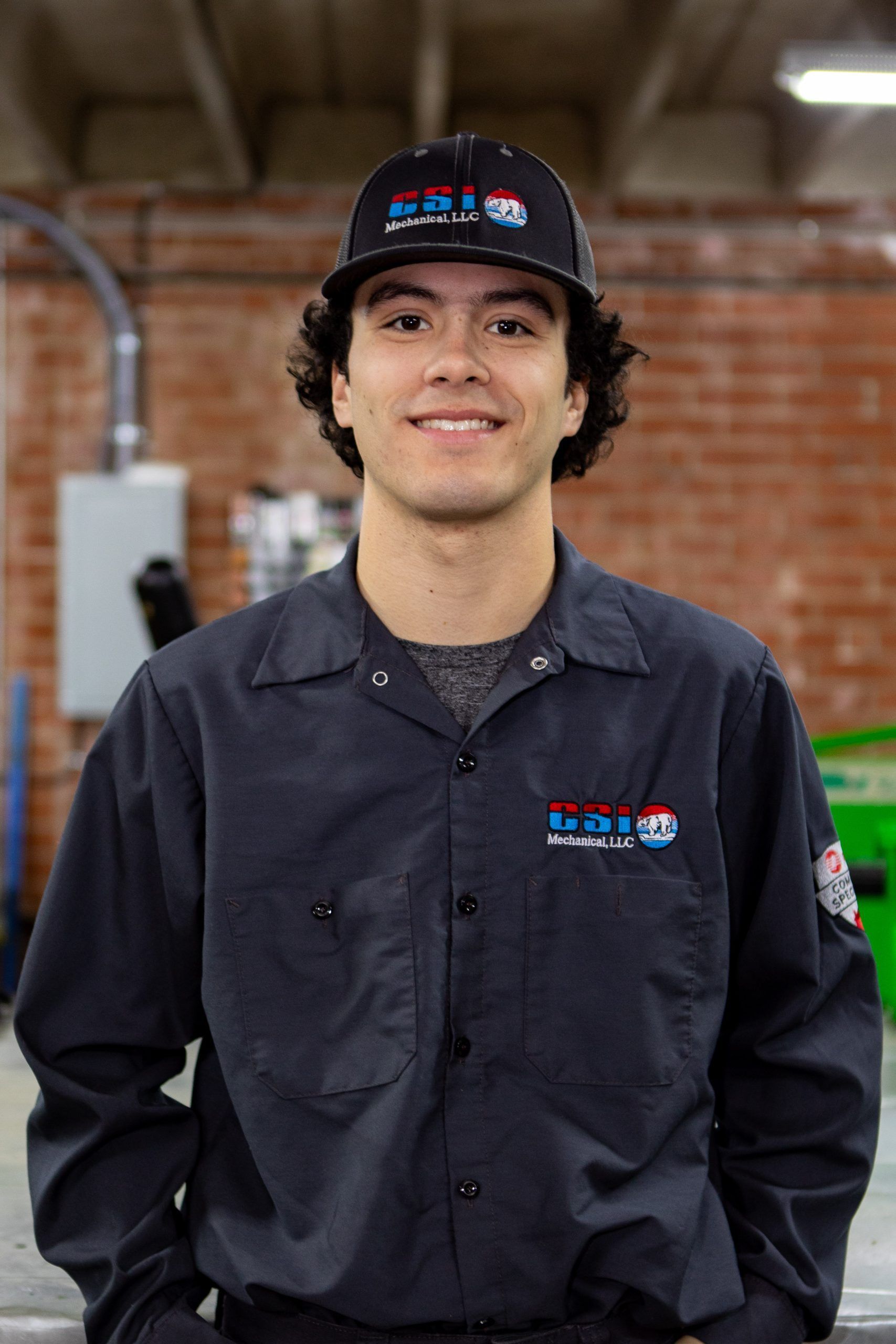 A person wears a dark work uniform and a branded baseball cap, smiling while standing before a brick wall in a workspace.