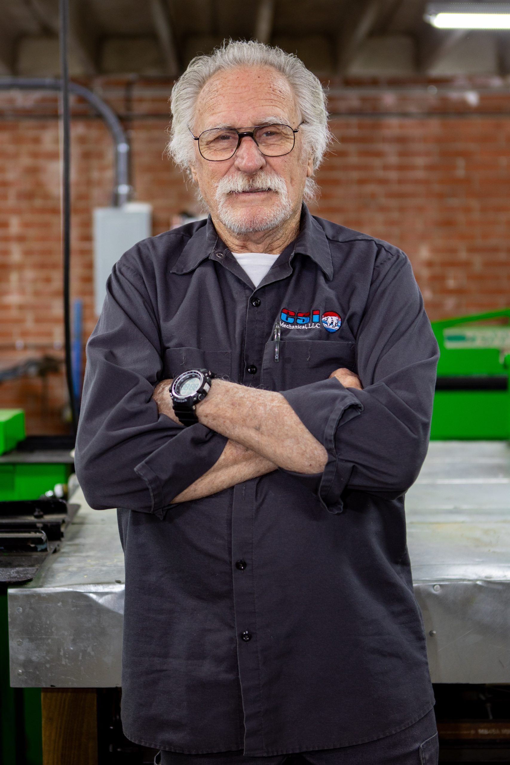 A person wearing glasses and a dark button-up shirt standing with arms crossed in a workspace with a brick wall background.