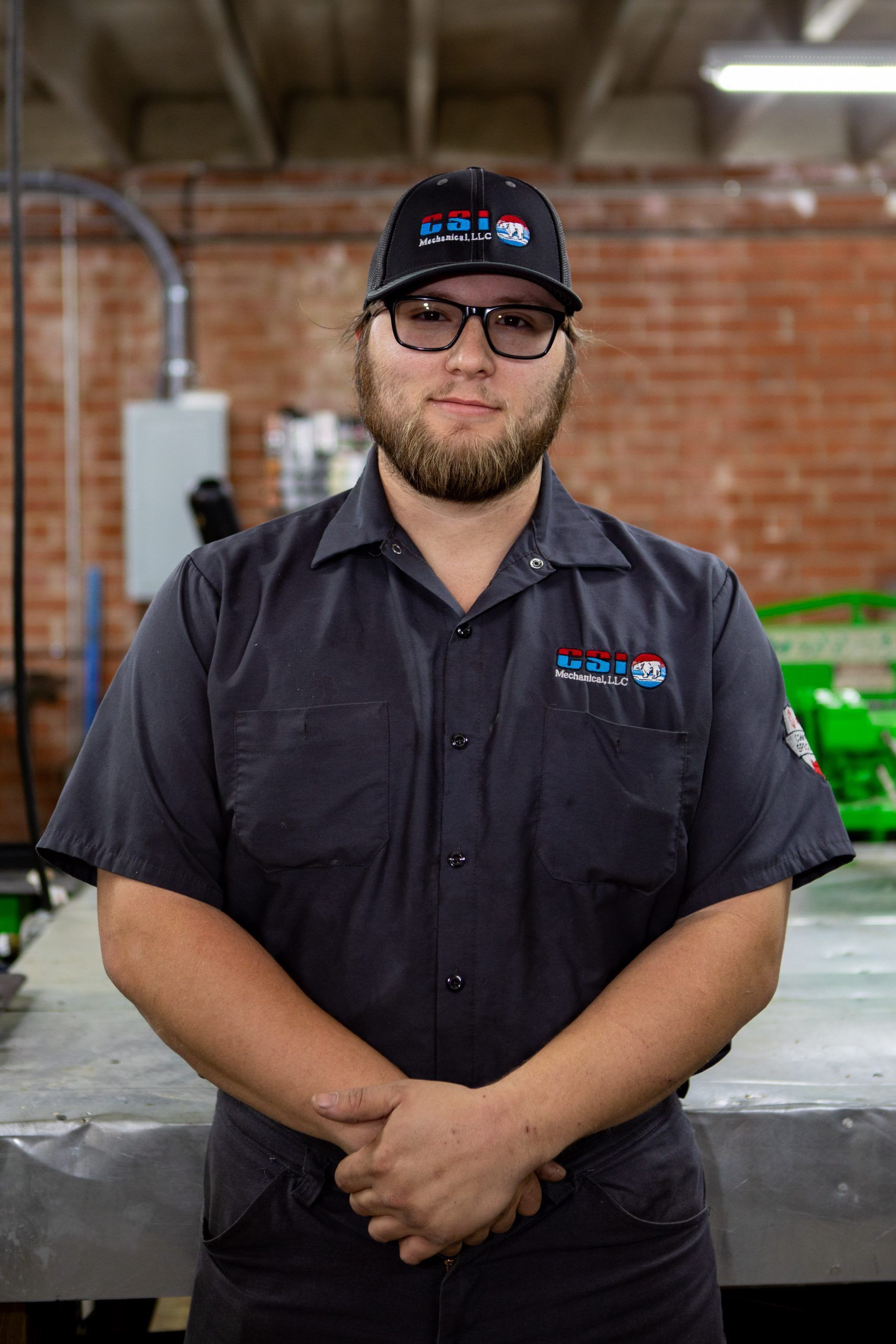 A person wearing a gray work shirt and branded cap standing with hands clasped in an industrial shop with brick walls.