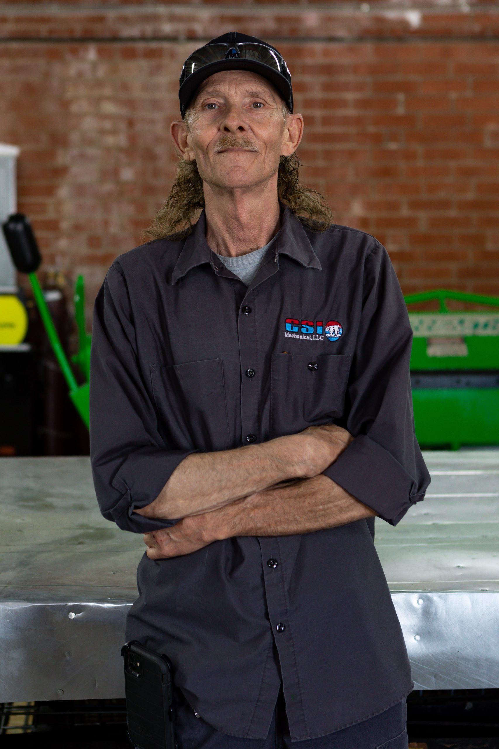 A person with curly hair wearing a dark shirt and cap, standing with arms crossed in a workshop with a brick wall.