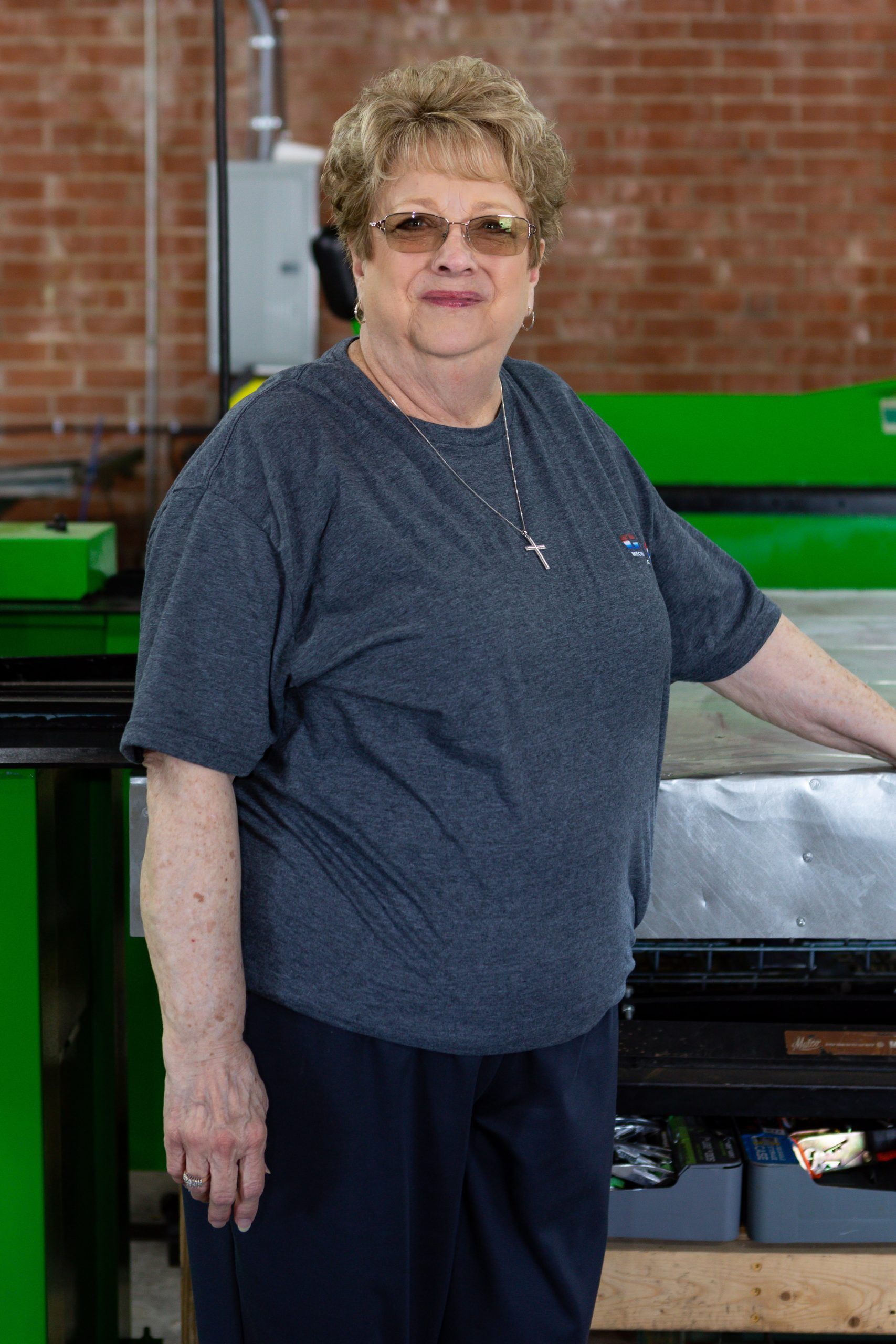A person wearing a grey t-shirt and glasses standing in a workspace with green industrial machinery and brick walls.