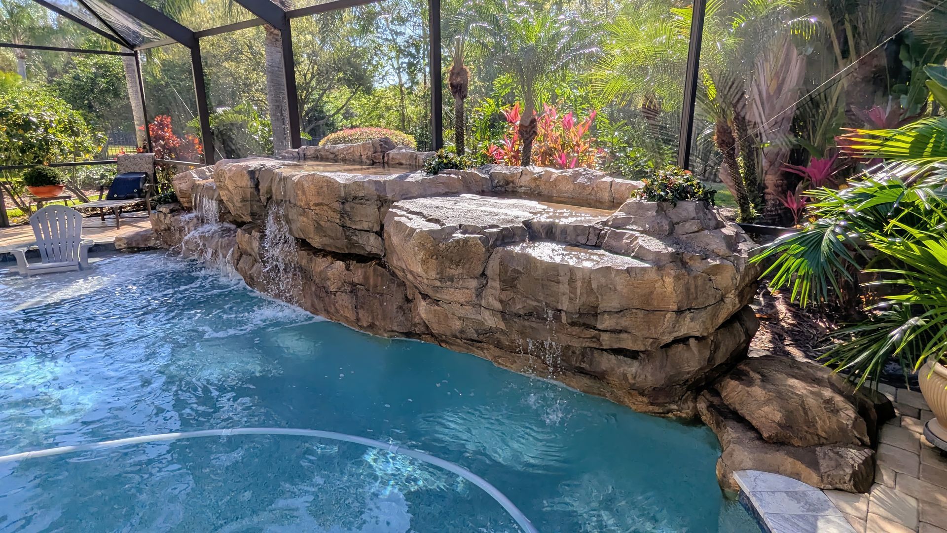 Backyard pool with a stone waterfall wall, blue water, and lush tropical plants under a screened enclosure
