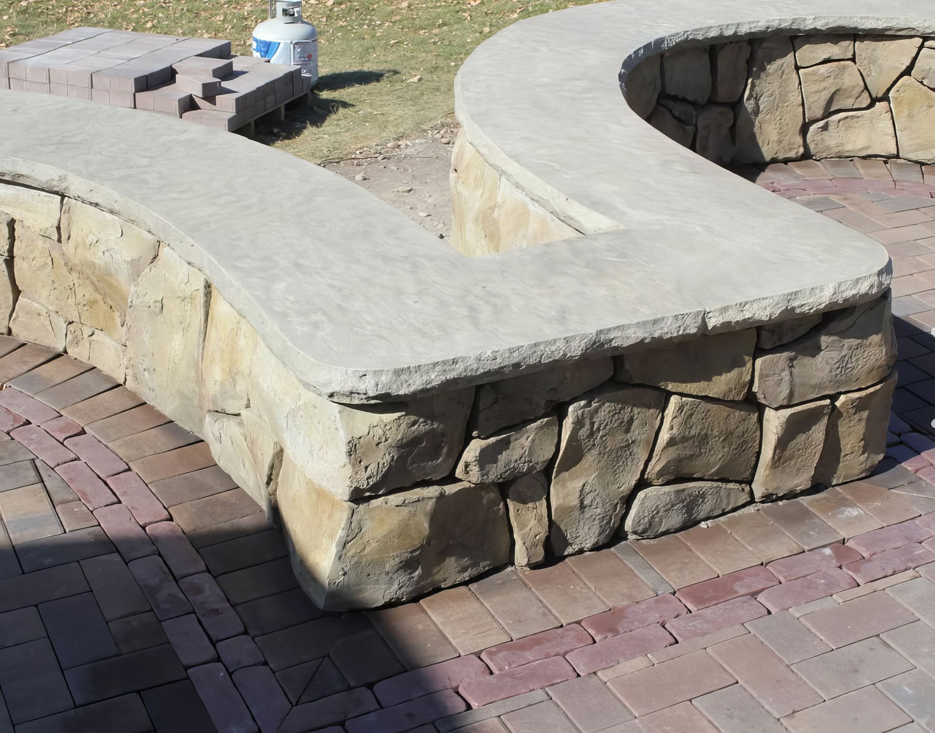 Curved stone bench with tan seat and stacked stone base beside a brick patio and fire pit area
