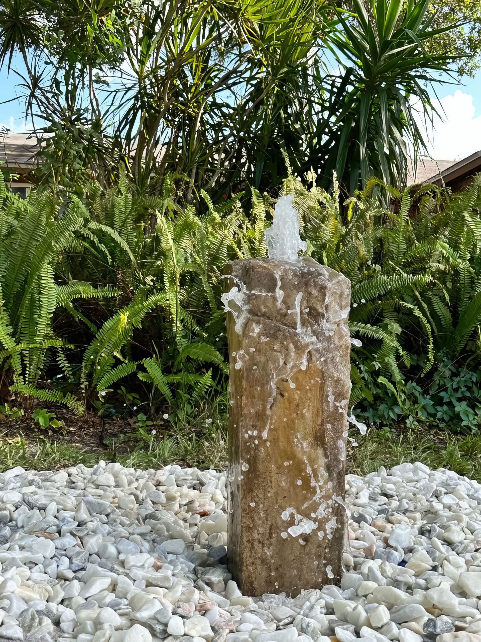 Stone fountain jet in a gravel garden with lush tropical plants in the background