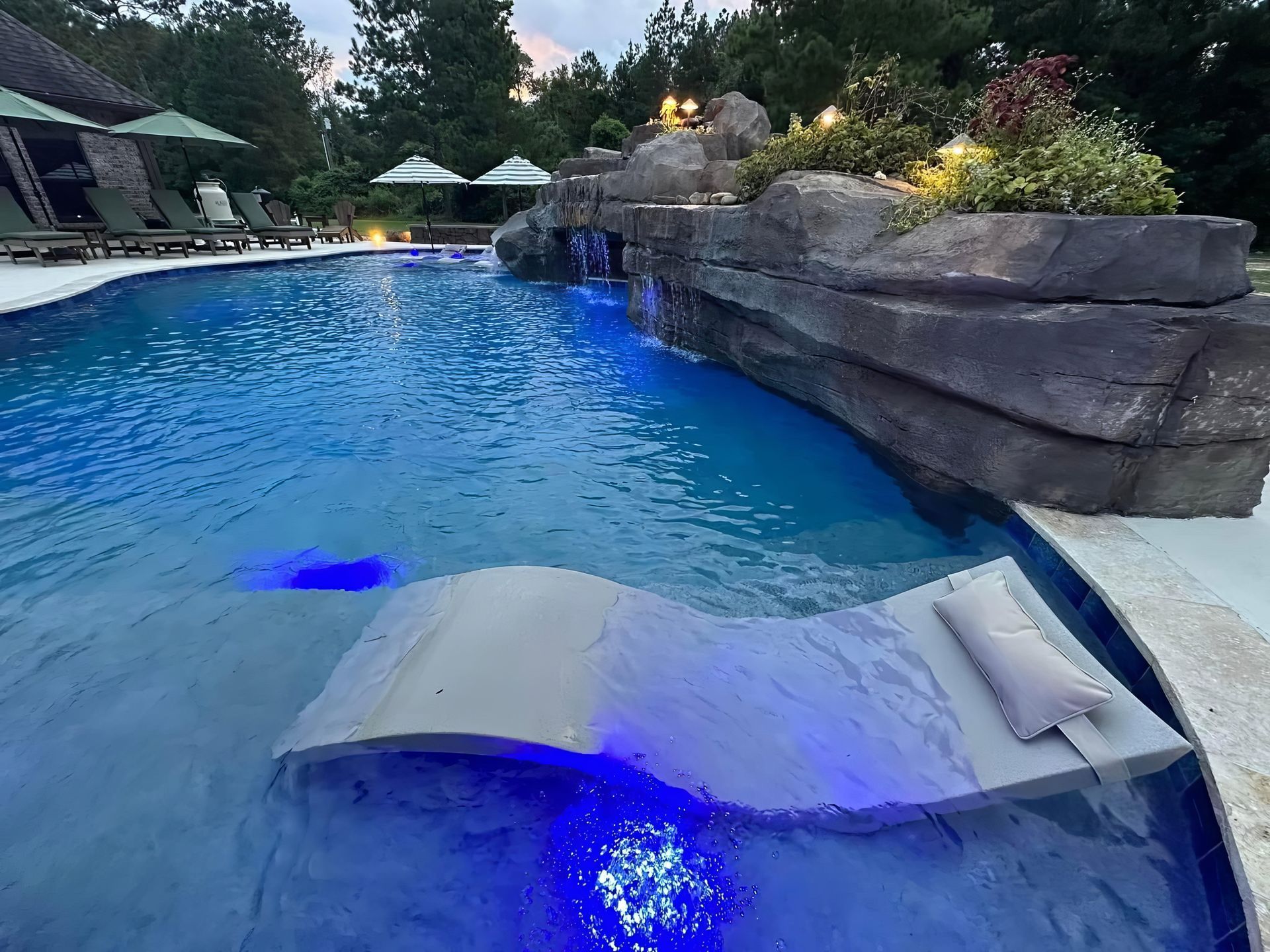 Blue-lit backyard pool with a curved stone waterfall and cushioned lounge seats on the water shelf