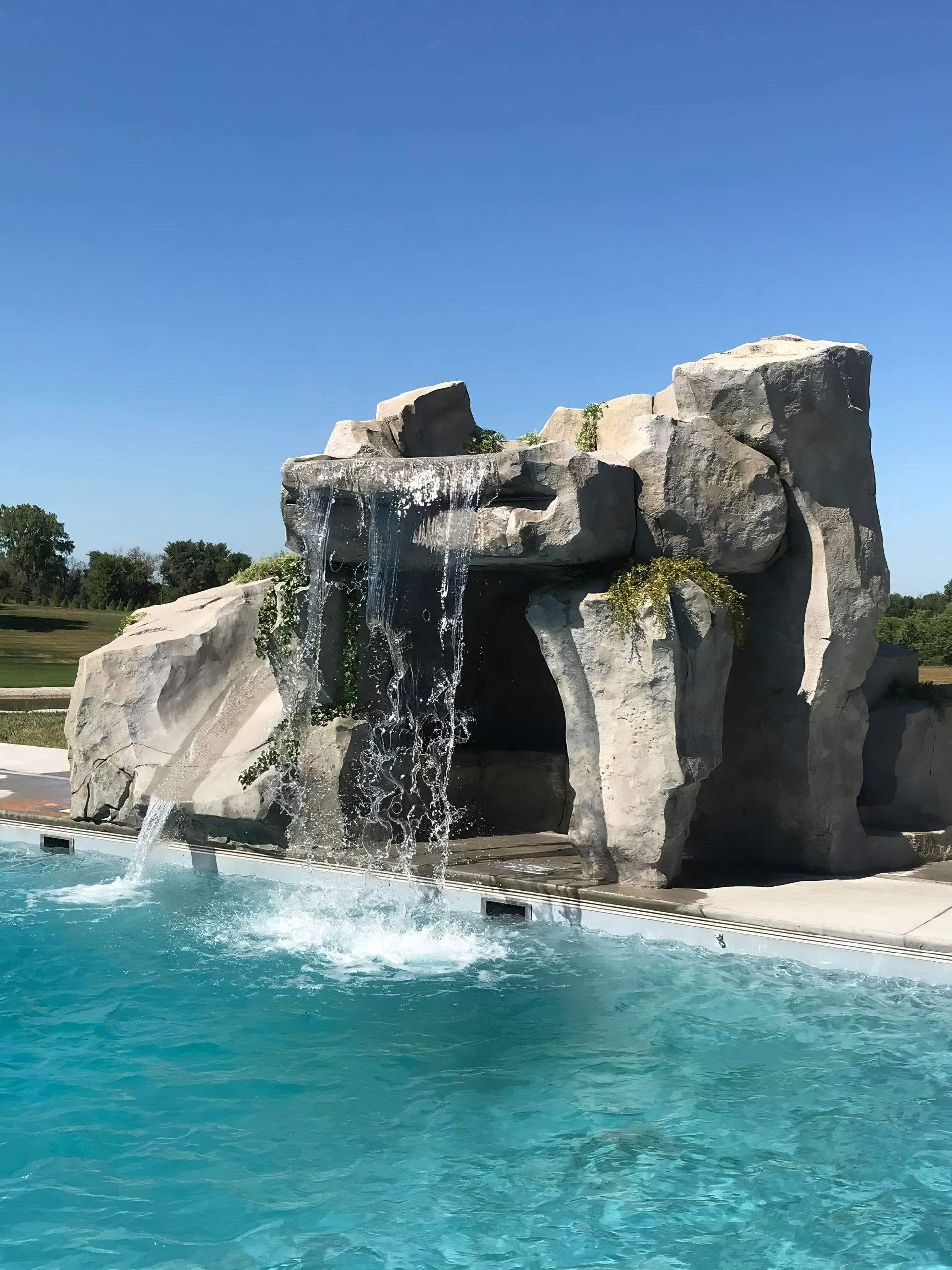 Rock waterfall spilling into a bright blue swimming pool under a clear sky