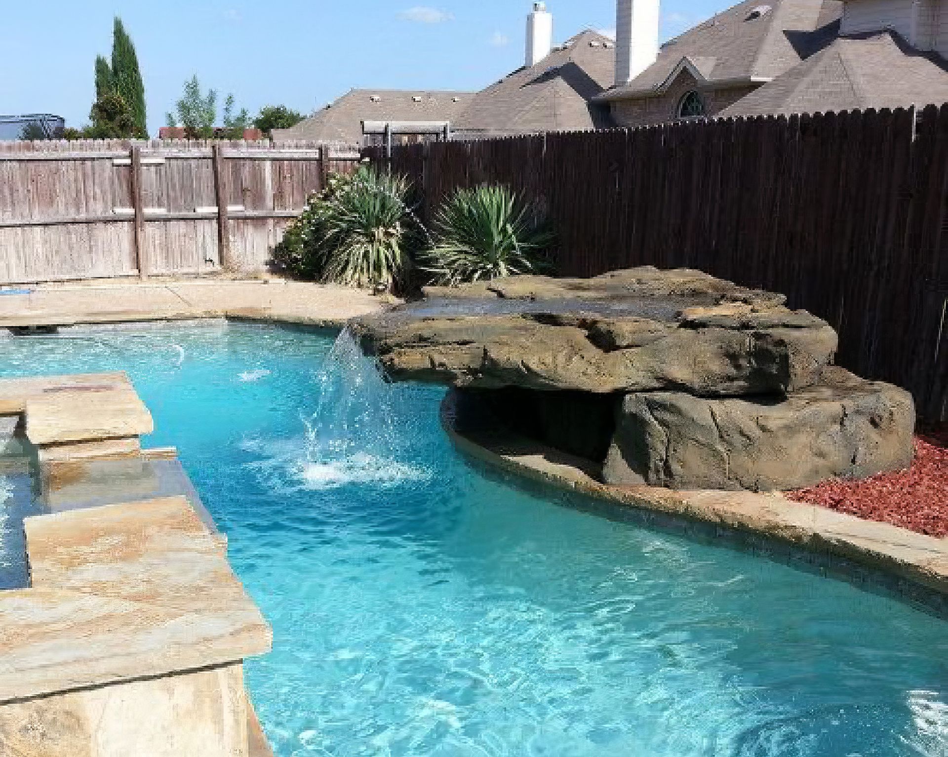 Backyard swimming pool with a rock waterfall and a wooden fence under a sunny sky
