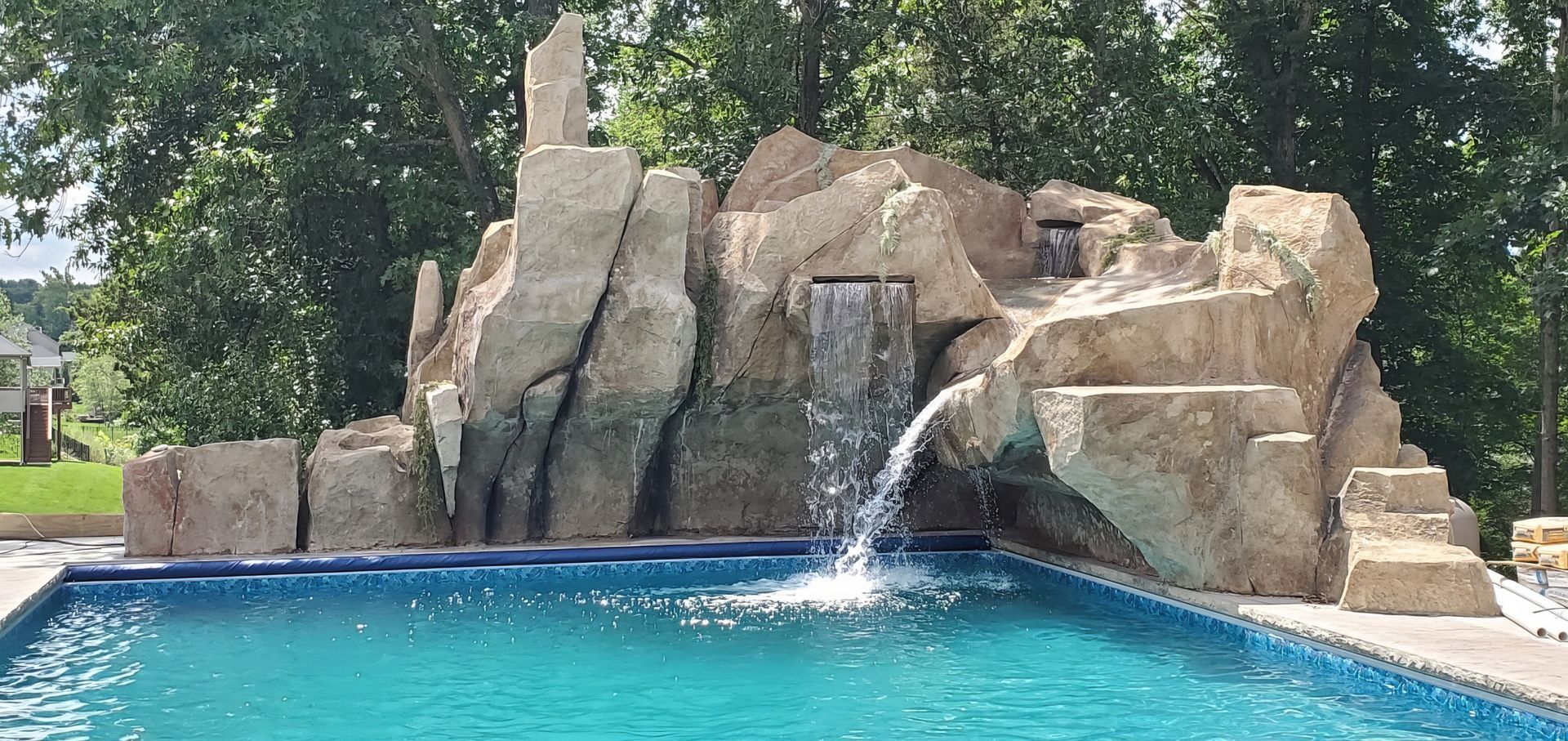 Rock waterfall spilling into a turquoise swimming pool surrounded by large tan rocks and trees