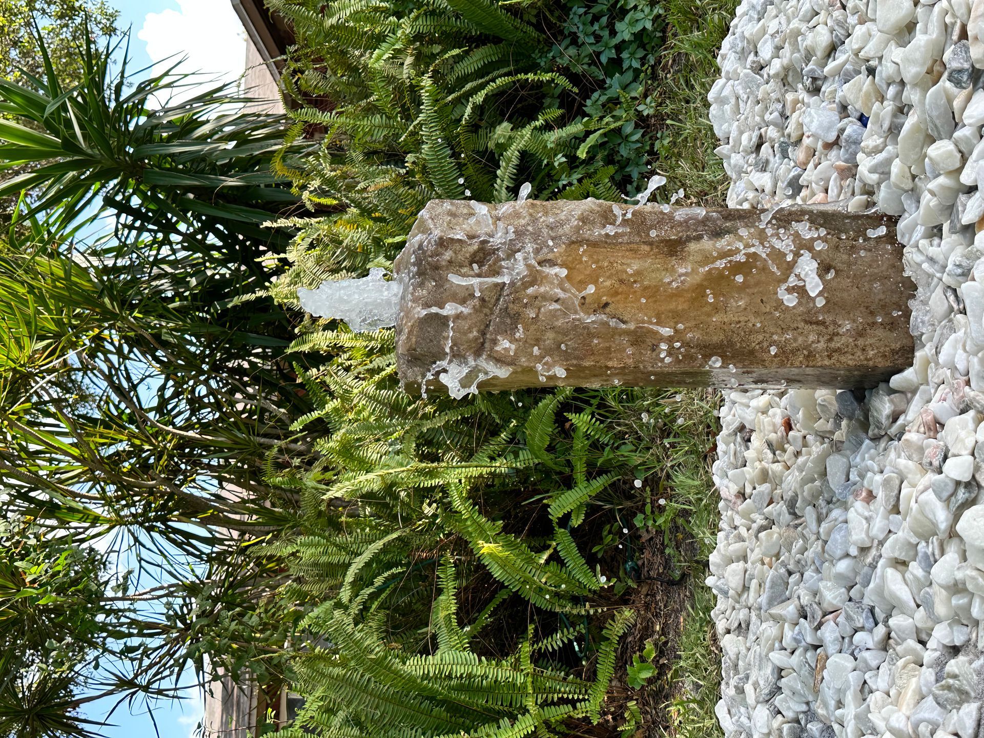 Stone fountain in a garden with water bubbling from a central pillar, surrounded by rocks and greenery