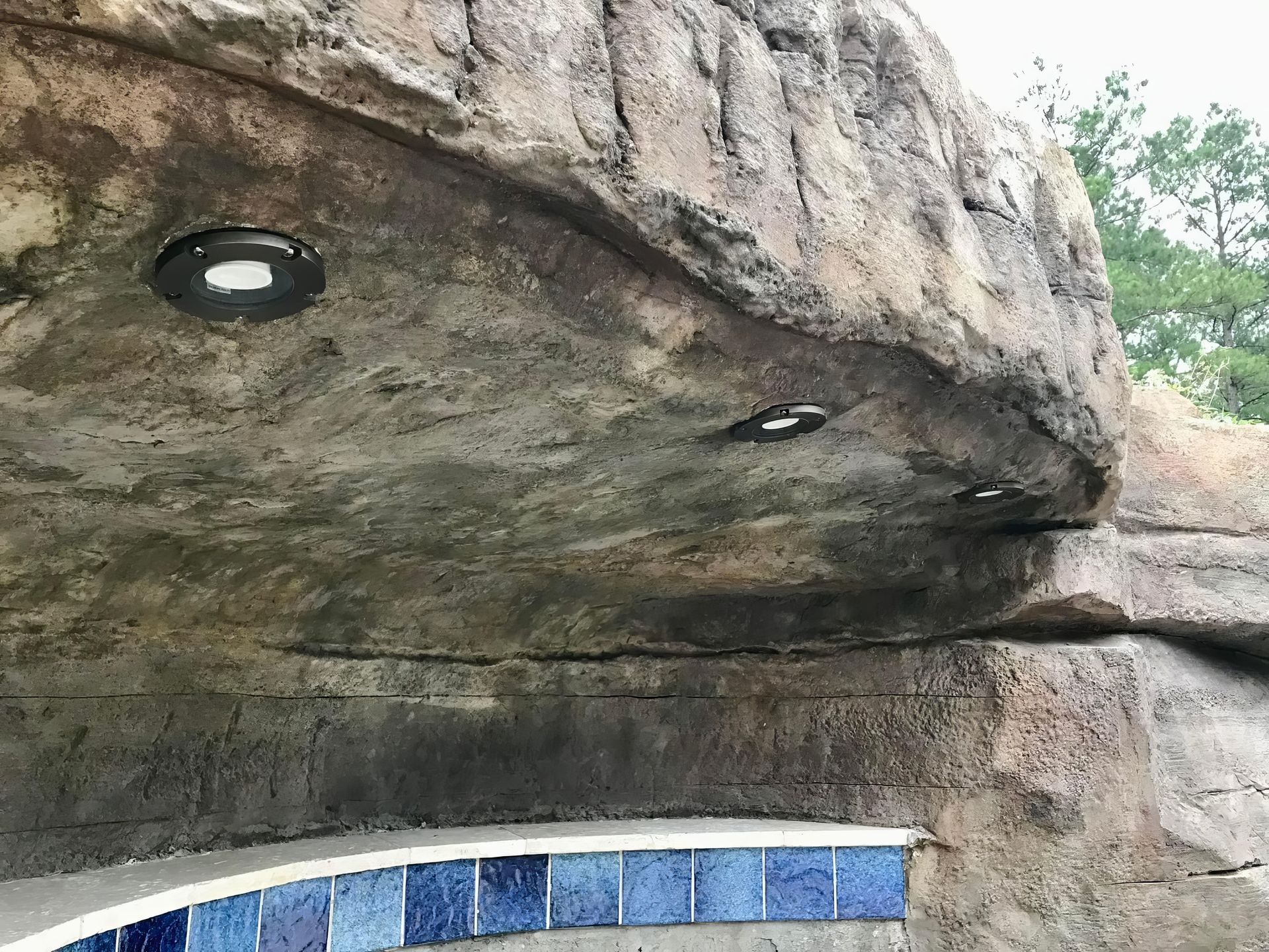 Rock overhang above a curved blue-and-white tiled pool edge with recessed lights and trees beyond.