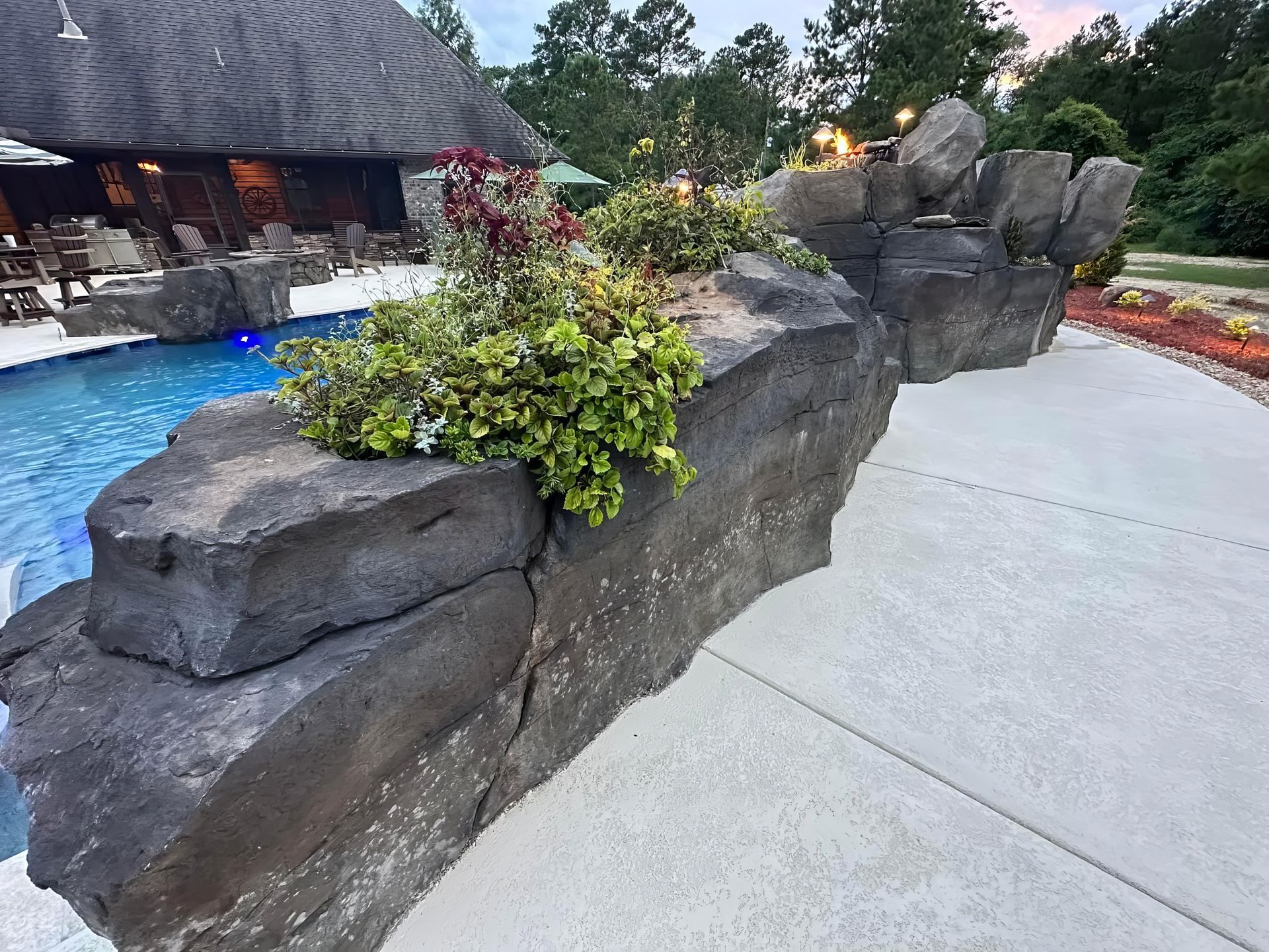Rock garden beside a concrete walkway, with a blue pool and thatched-roof building in the background.