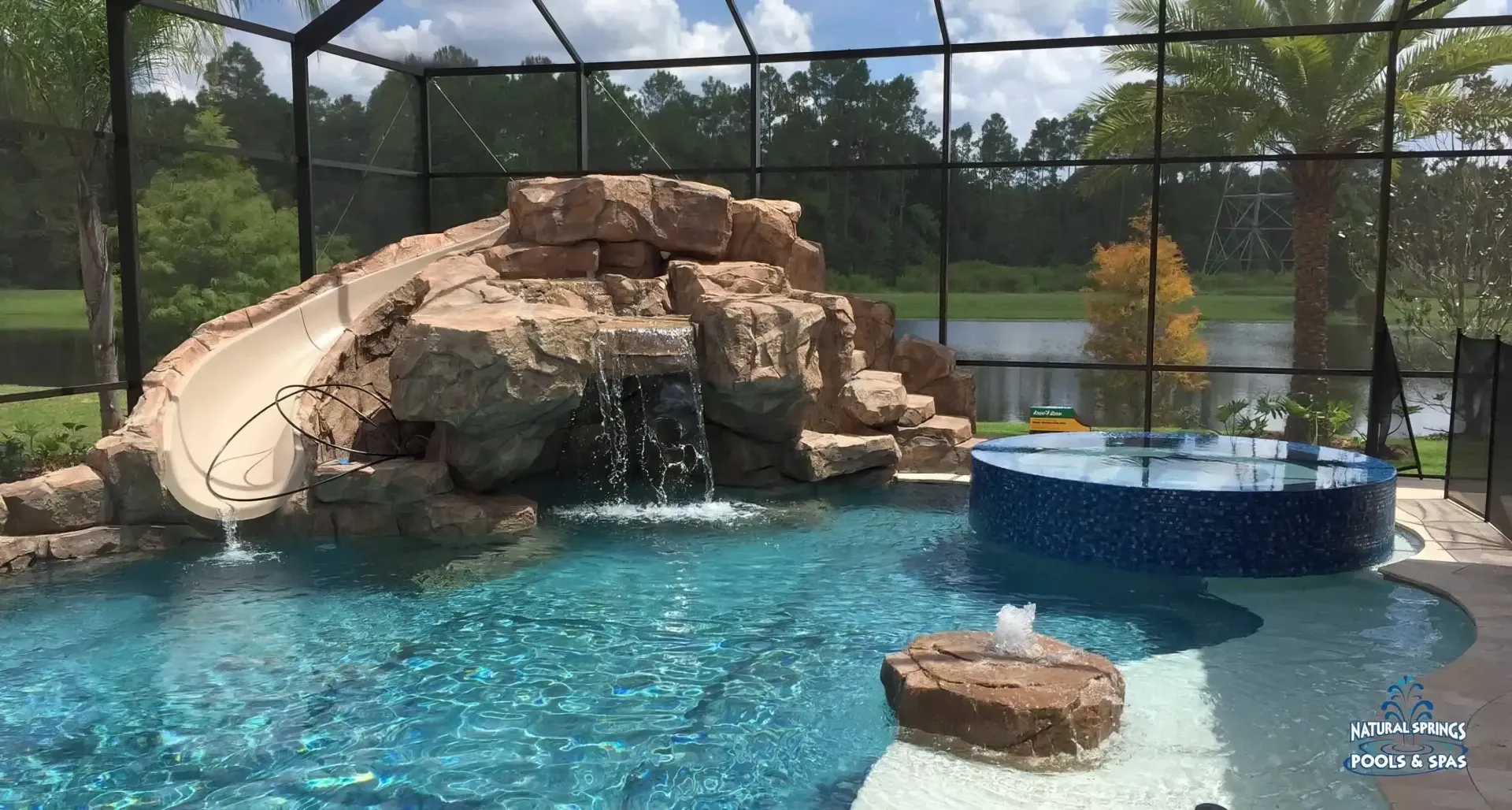 Outdoor zoo pool with rocks, a tan slide, and a round blue platform behind a safety fence.