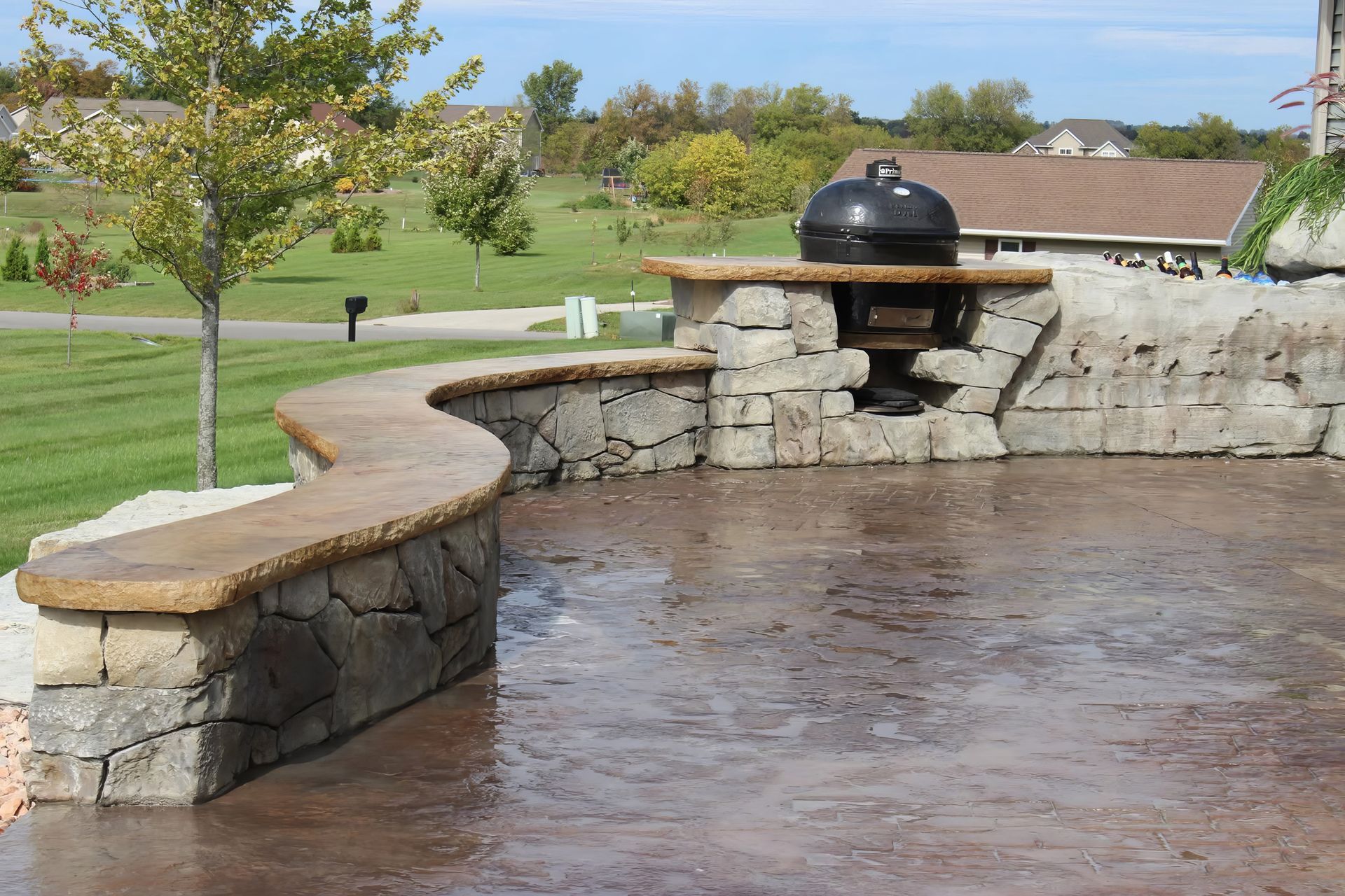 Curved stone patio with a built-in black grill overlooking a green park and trees