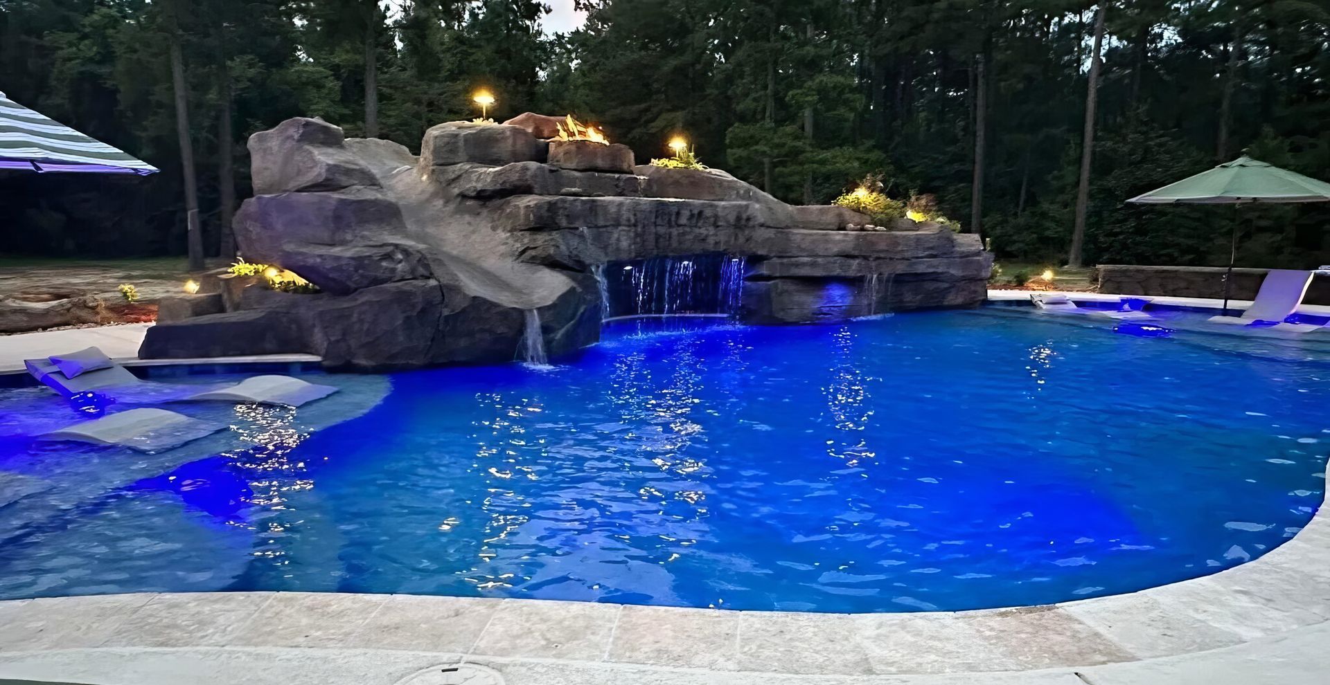 Backyard pool with blue lit water, rock waterfall, and patio seating at dusk