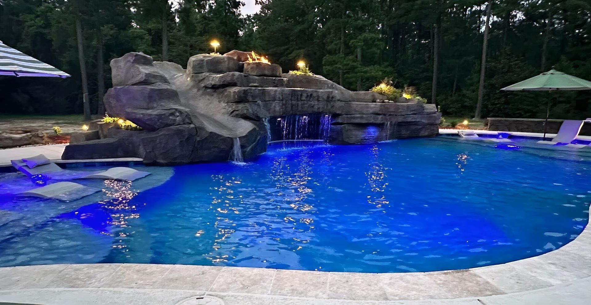 Blue-lit backyard pool with rock waterfall at dusk, surrounded by trees and lounge chairs
