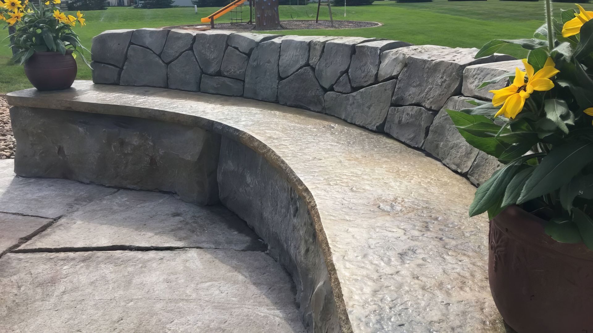 Curved stone bench and path in a garden with potted yellow flowers and stone wall backdrop