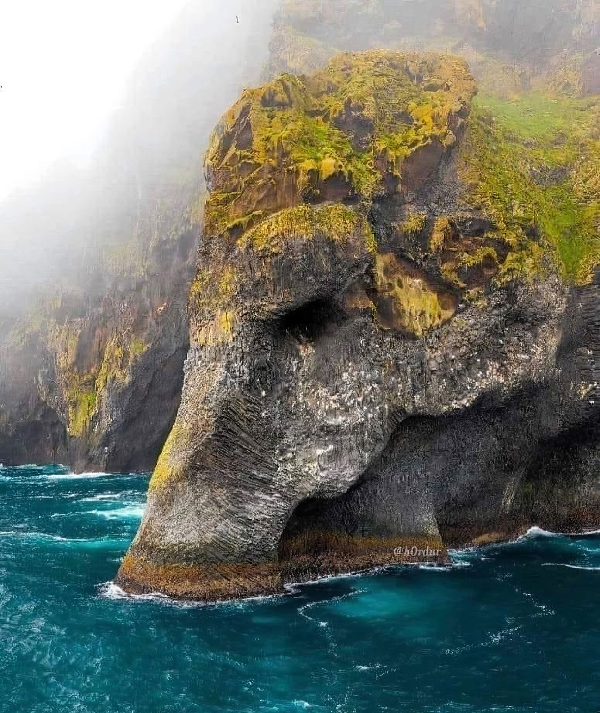 Foggy coastal cliff with yellow moss above turquoise ocean waves