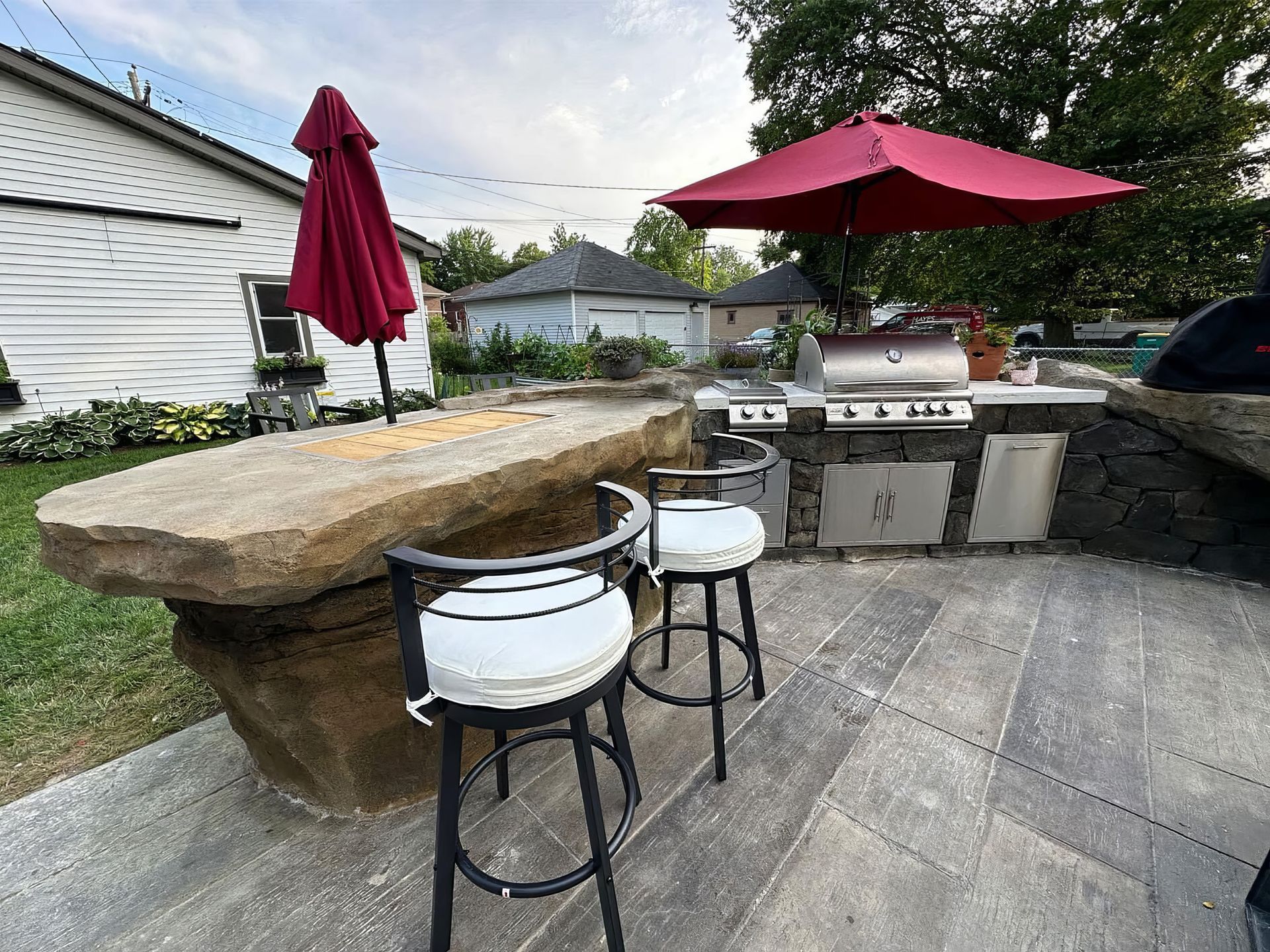 Backyard patio with stone bar, grill, red umbrellas, and two stools on a paved deck