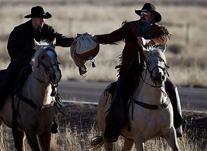 Two men are riding horses and one is holding a bag