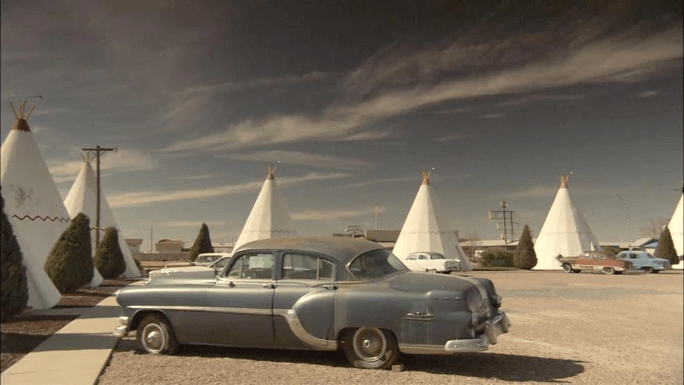 An old car is parked in a parking lot with teepees in the background