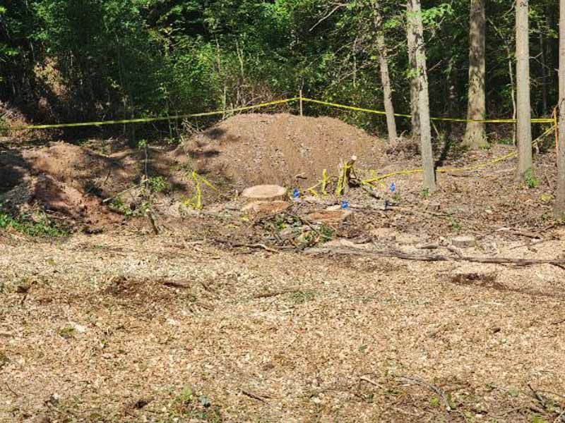 A construction site in a wooded area with a mound of excavated dirt, a round cover, and yellow caution tape.
