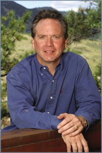 Man in blue shirt leans on a railing, smiling. Outdoor setting with trees.