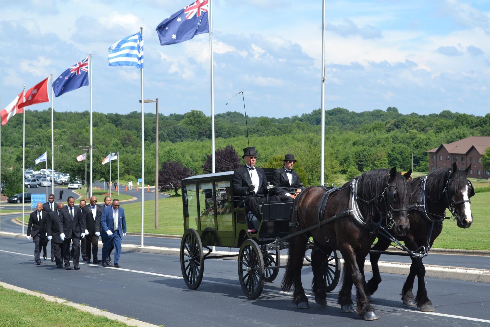 A horse-drawn hearse procession with people in suits, flags in the background.