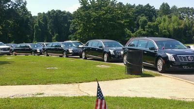A line of black hearses parked on green grass near trees and an American flag.