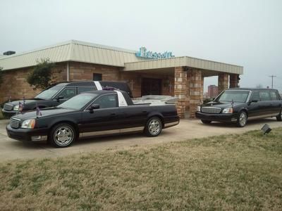 Three black Cadillac limousines parked outside a building with a blue sign, on a cloudy day.