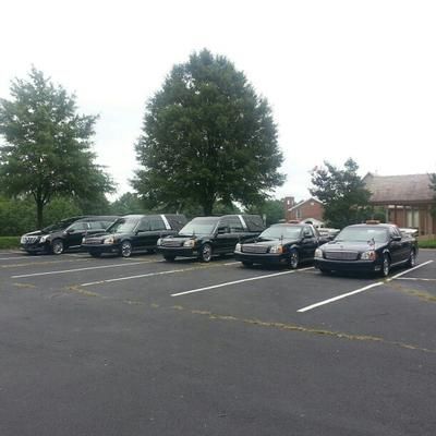 Six black hearses parked in a lot, near a building and trees.
