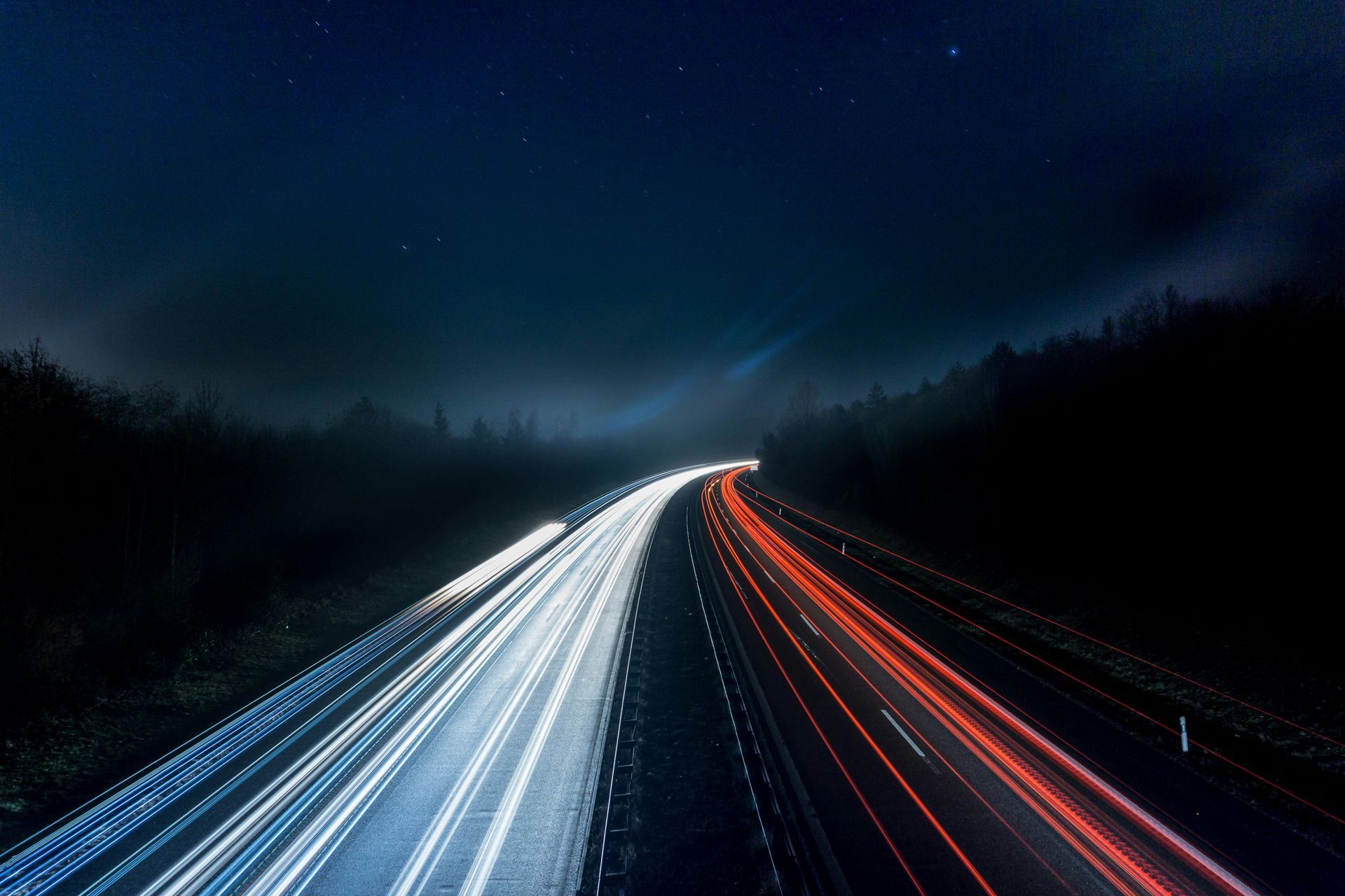 Long exposure of highway at night; streaks of white and red car lights on dark road, forest in background.