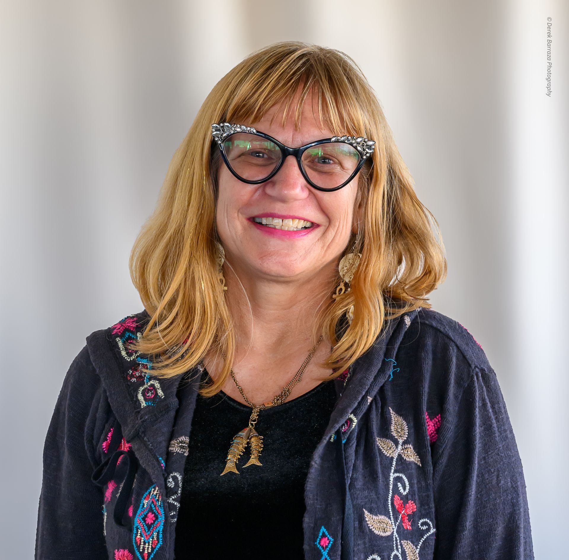 Woman with blonde hair, glasses, smiling, wearing a jacket with floral embroidery, and a necklace.