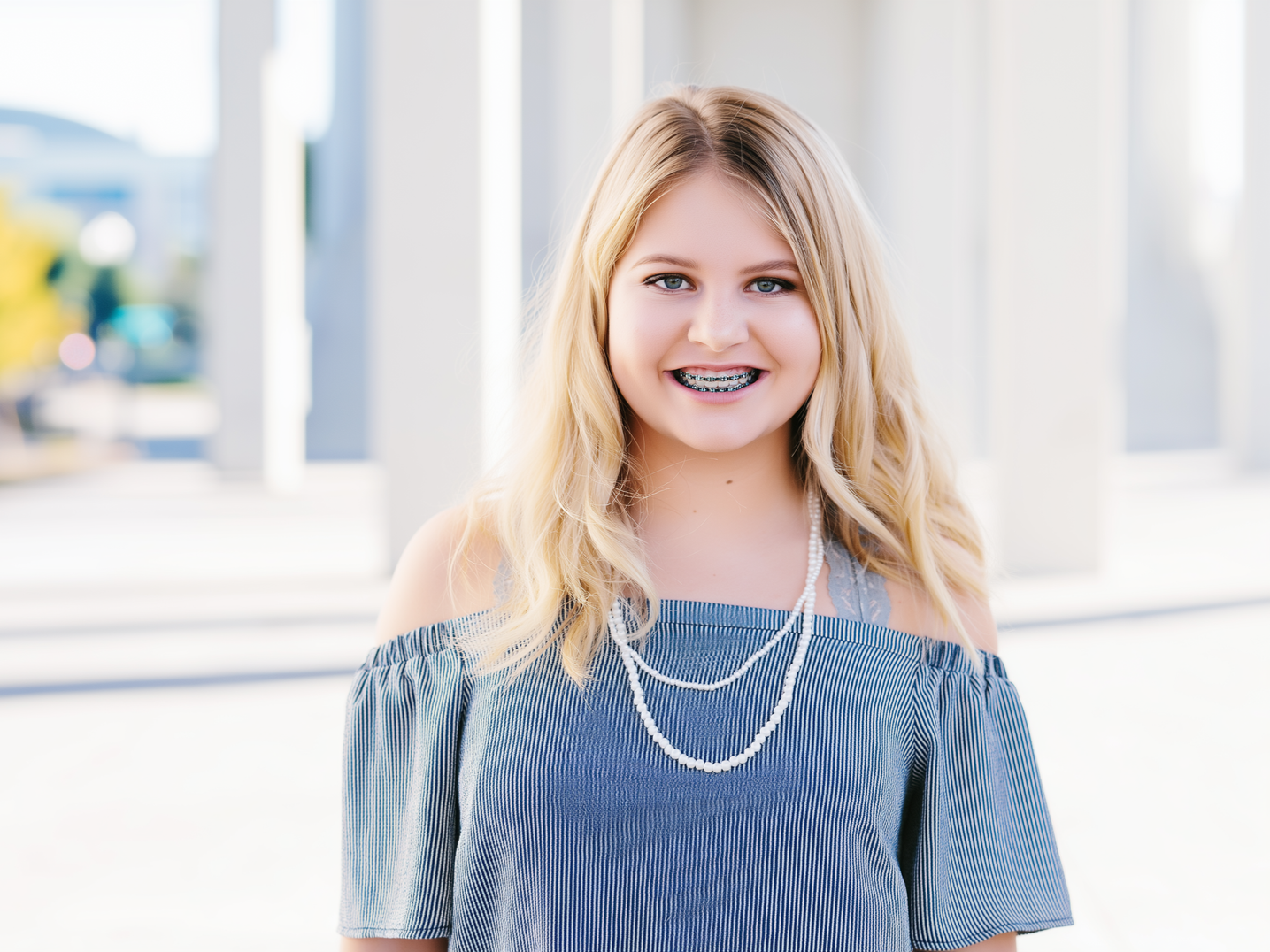 Blonde girl with braces wearing off-the-shoulder striped shirt and pearl necklace, smiling outdoors near white columns.