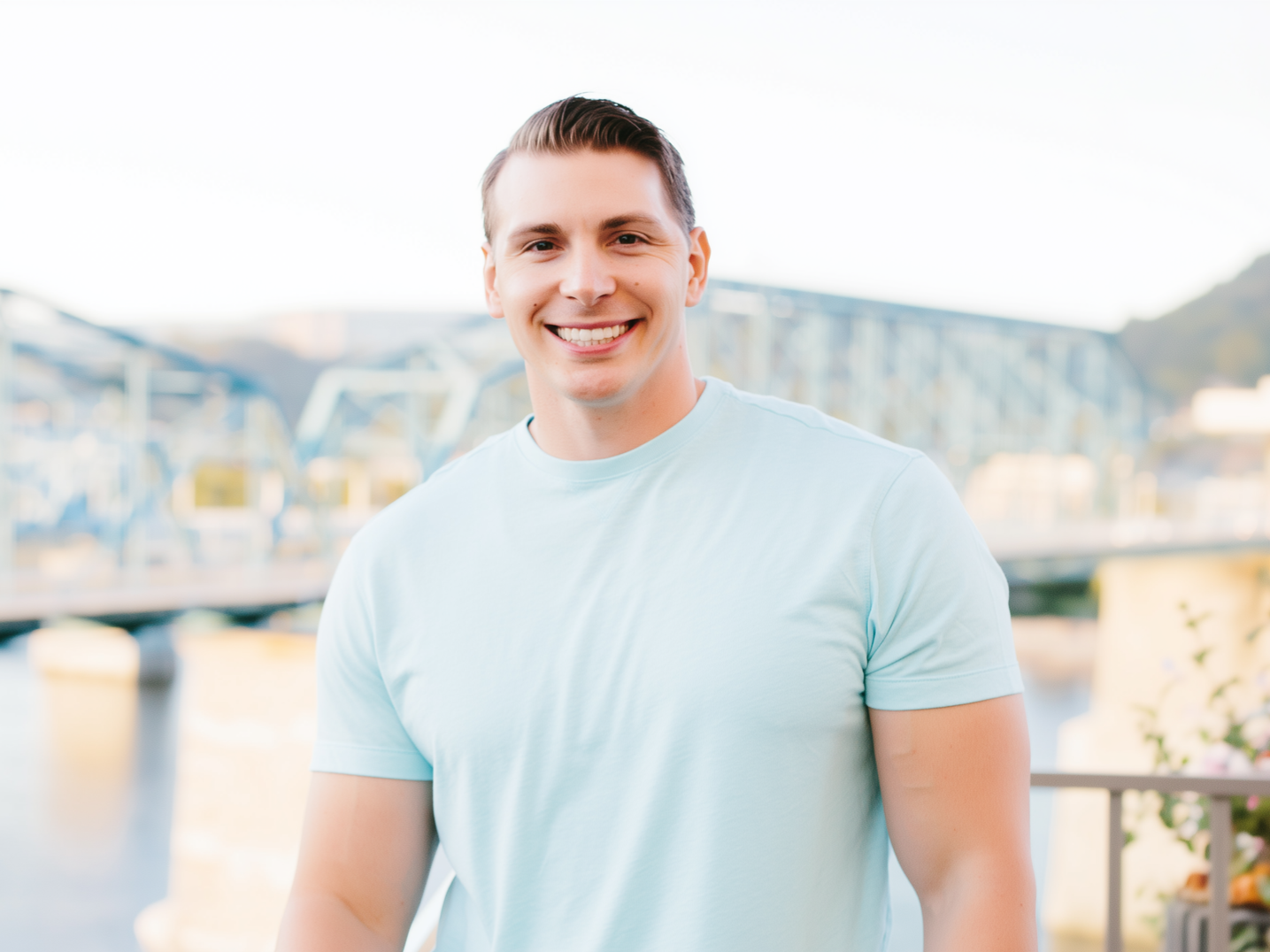 Smiling man in a light blue shirt standing outdoors with a blurred waterfront cityscape behind him