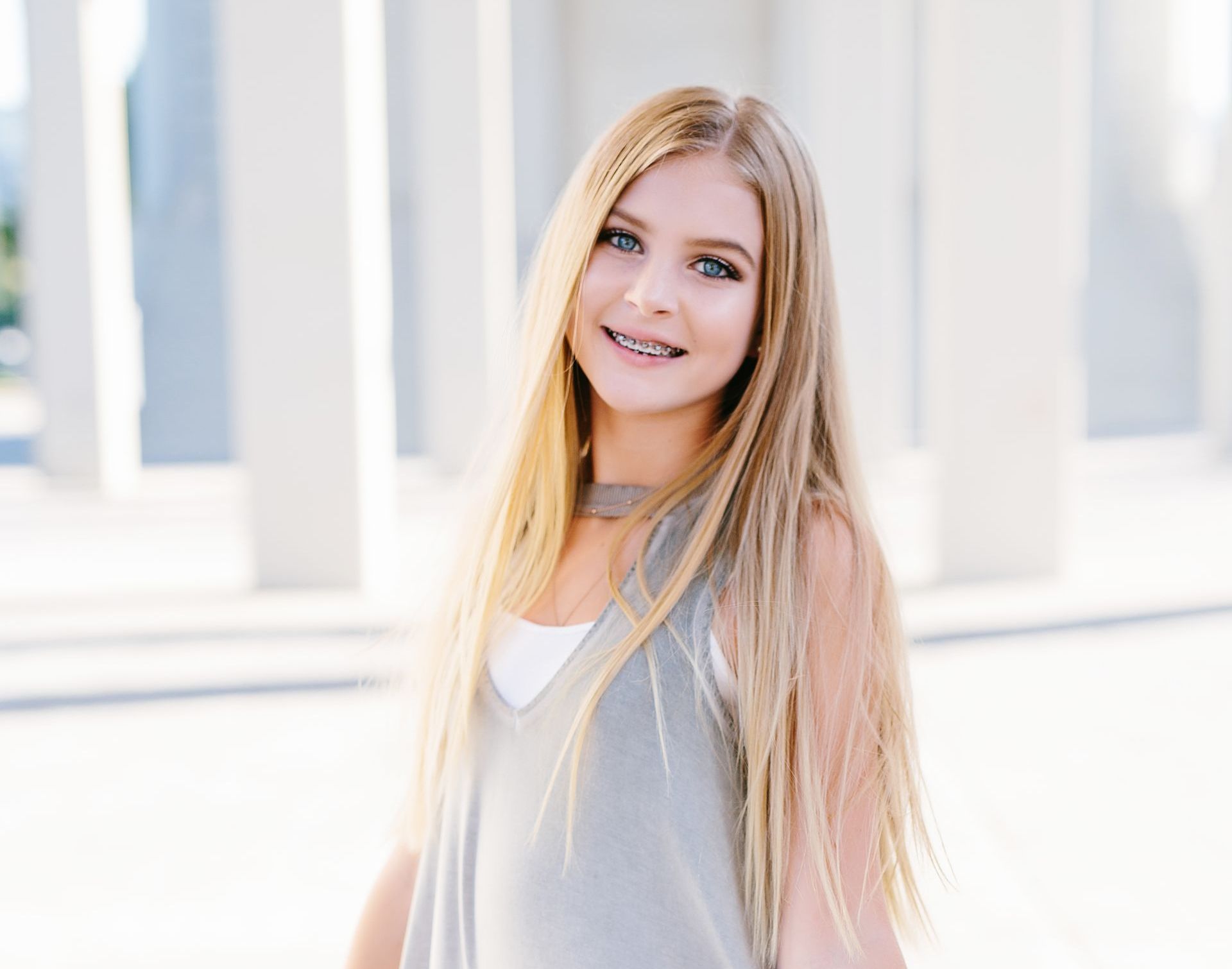 Blonde teenager with braces smiles in front of a white building. She wears a gray tank top and a choker.
