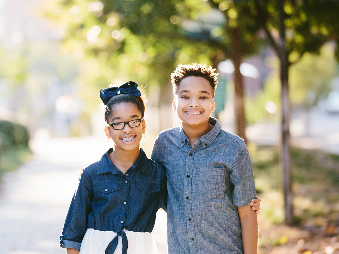 Two smiling children, a girl with glasses and a bow, and a boy with his arm around her, outside.
