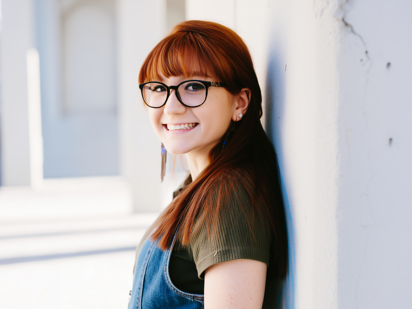 A person with red hair and glasses, wearing a denim overall, smiling while leaning against a white pillar.