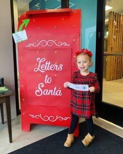 A little girl is standing in front of a mailbox that says `` letters to santa ''.