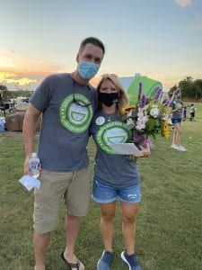 A man and a woman wearing masks are standing next to each other in a field holding flowers.