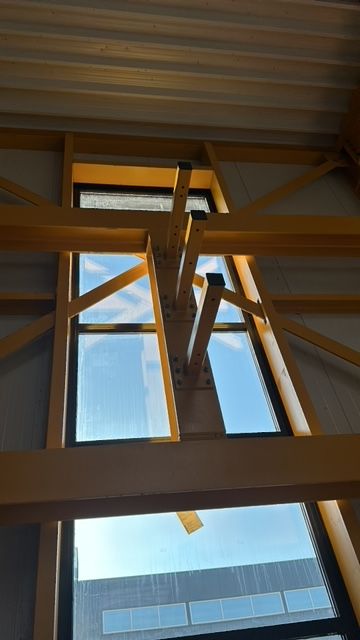 Interieurbeeld van hoge ramen omlijst door gele stalen balken tegen een lichtblauwe hemel.