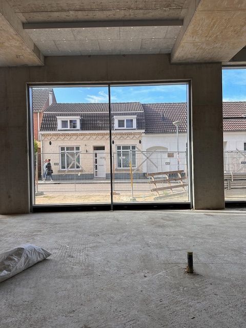 Uitzicht door een groot raam van een gebouw in aanbouw, met zicht op een straat met huizen en een voetganger.