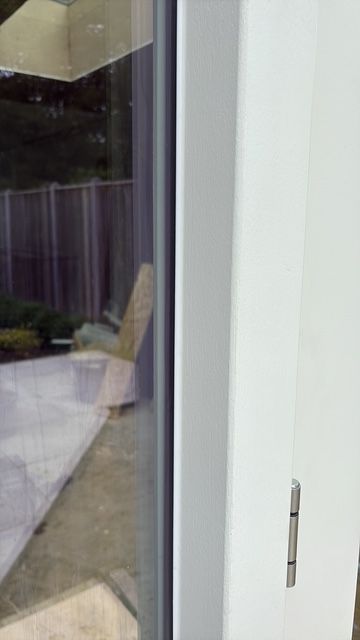 Close-up van een wit deurkozijn en scharnier, met een wazig zicht op een terras door de glasplaat.