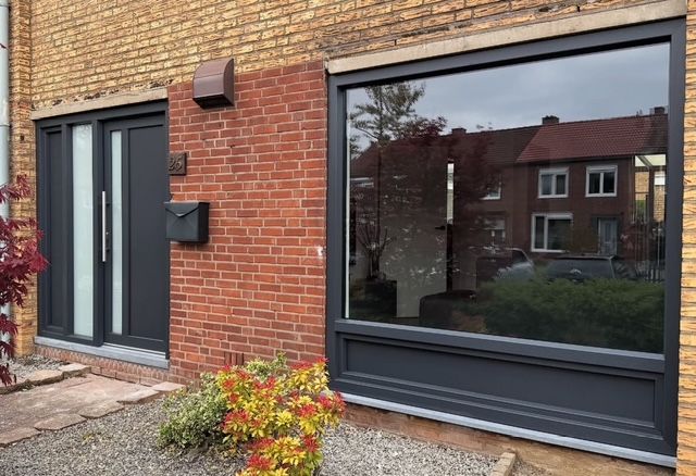 Donkergrijze deur- en raamkozijnen in een bakstenen gebouw; weerspiegeling van huizen in het raam.