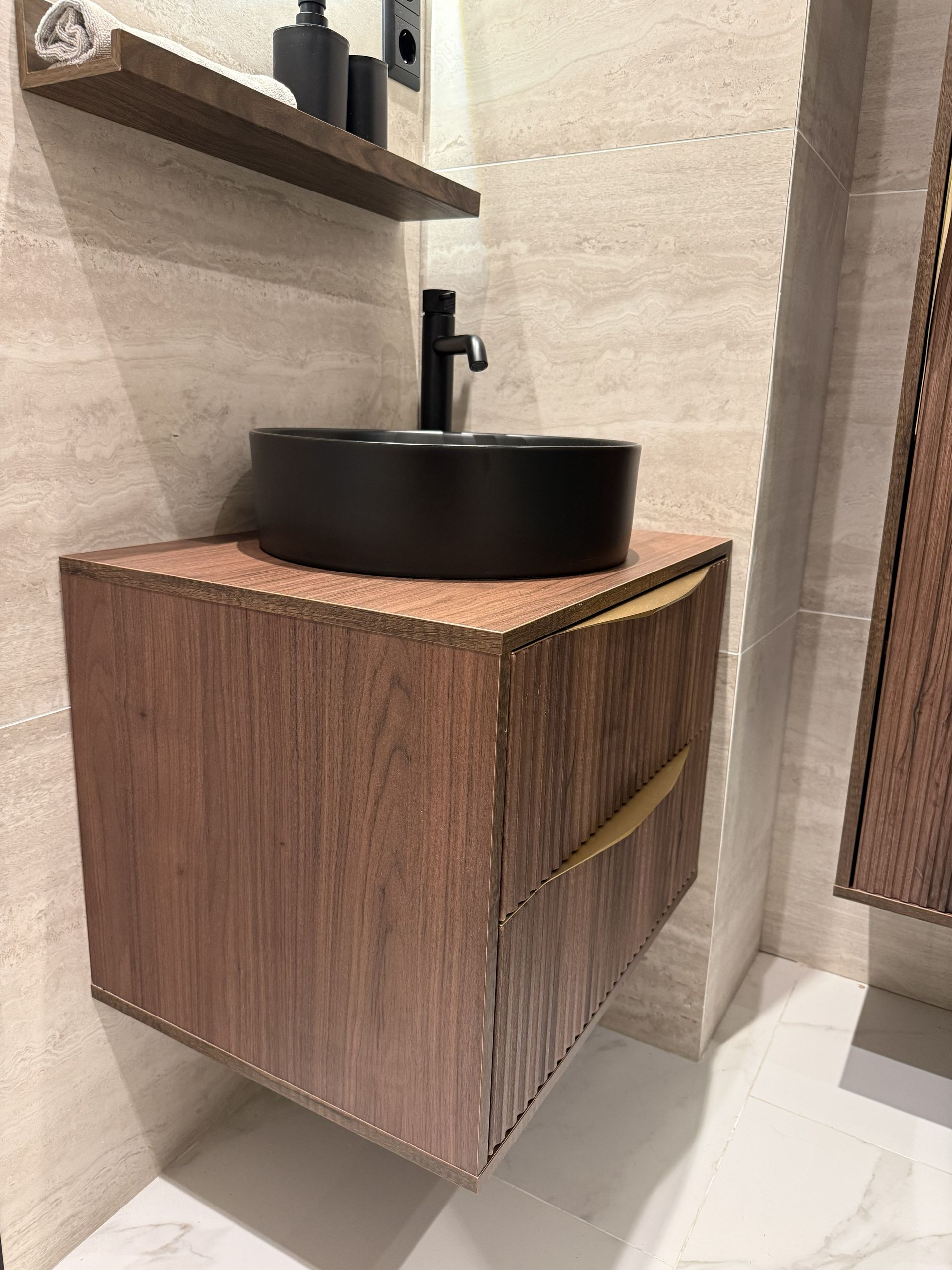 Een modern, zwevend houten badkamermeubel met een zwarte opbouwspoelbak en kraan tegen een lichtgekleurde stenen tegelwand.