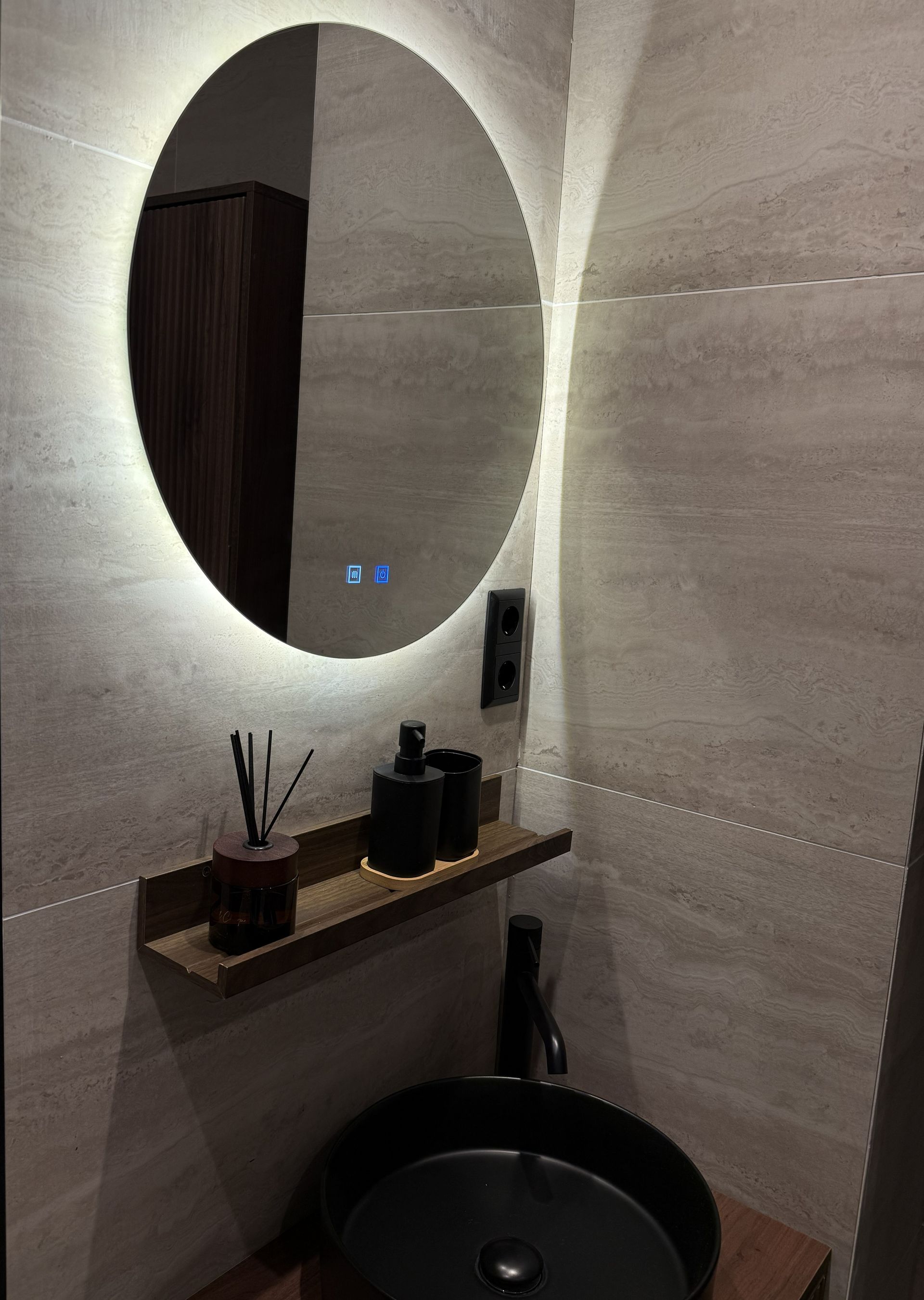 Een wastafel in de badkamer met een zwarte opbouwkom, een klein plankje en een ronde spiegel met achtergrondverlichting, tegen een muur van lichtgekleurde tegels.