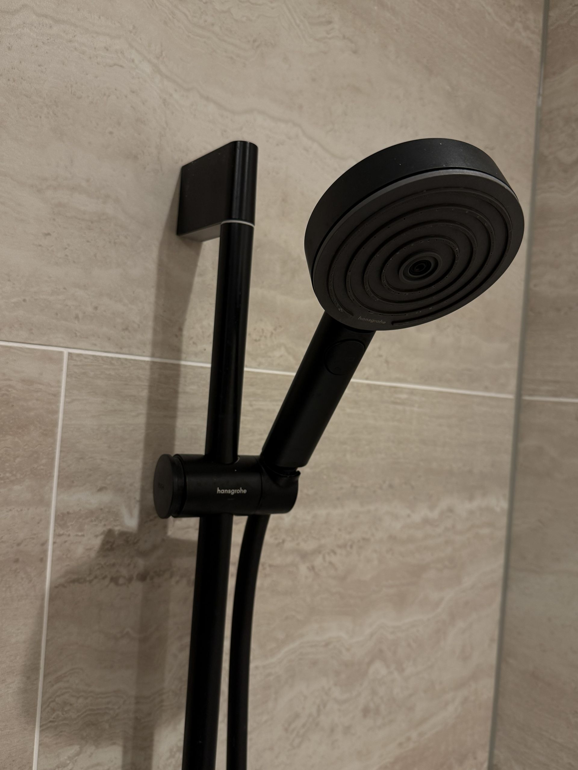 Een zwarte handdouchekop, bevestigd aan een verticale schuifstang, gemonteerd tegen een lichtbeige betegelde douchewand.