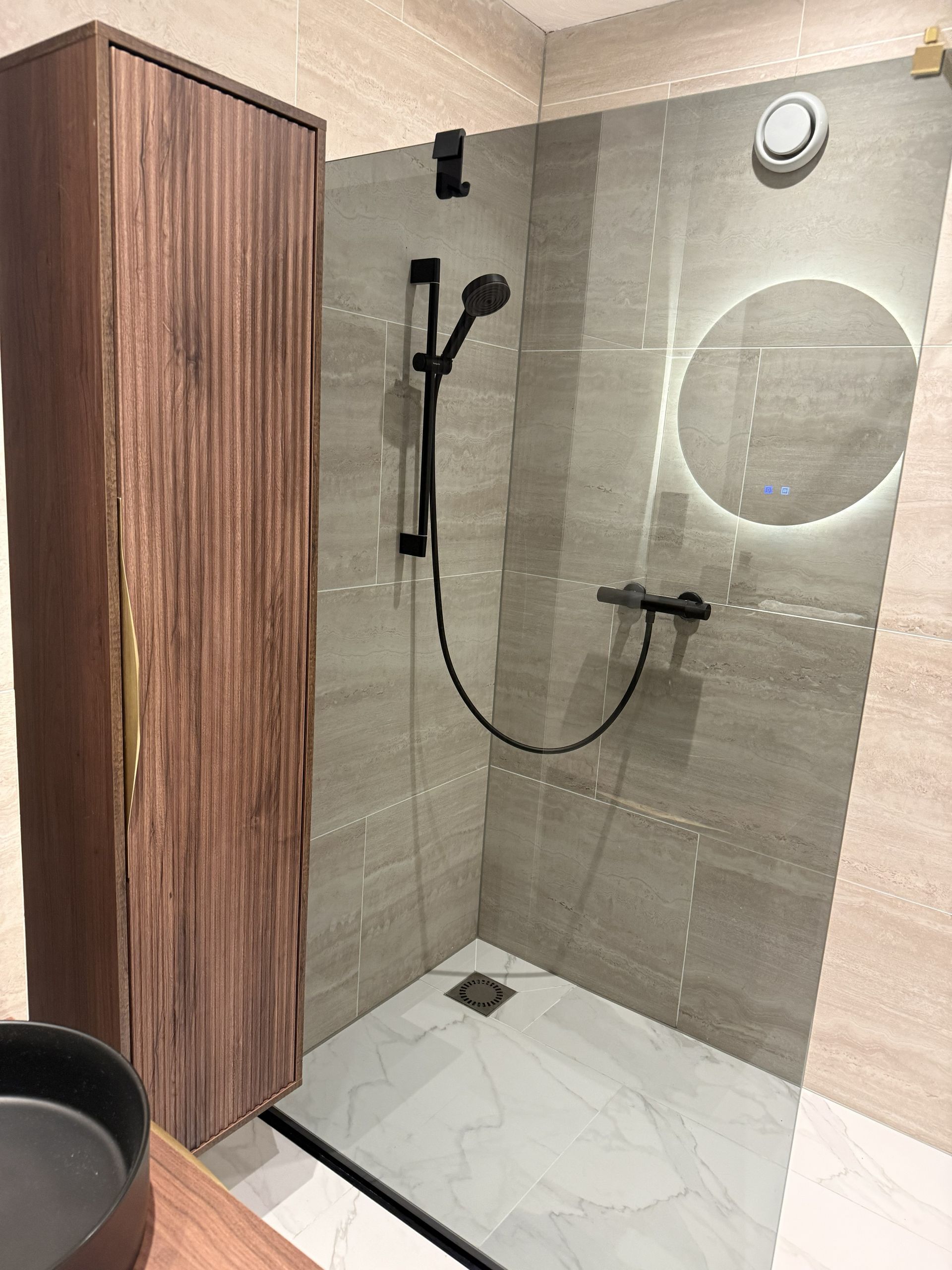 Een moderne douche met bruine tegelwanden, een zwarte handdouche, een ronde spiegel met achtergrondverlichting en een houten kast.