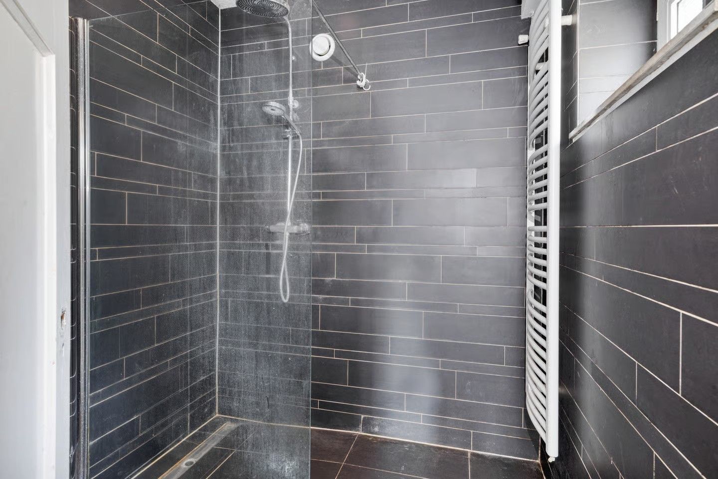 Een inloopdouche met een glazen wand, donkergrijze tegelwanden, een aan de muur gemonteerde douchekop en een witte handdoekradiator.