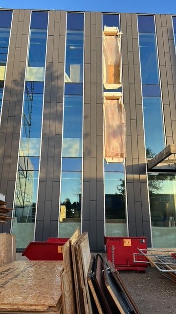 De buitenkant van het gebouw heeft donkere panelen en hoge ramen; eronder bevinden zich bouwmaterialen en een afvalcontainer.